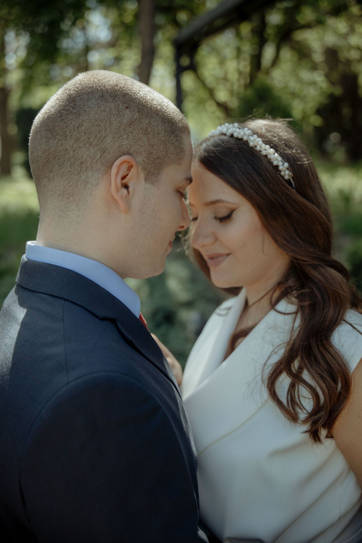 A bride and groom share an intimate forehead touch moment outdoors, with the bride wearing a white dress and pearl headband.