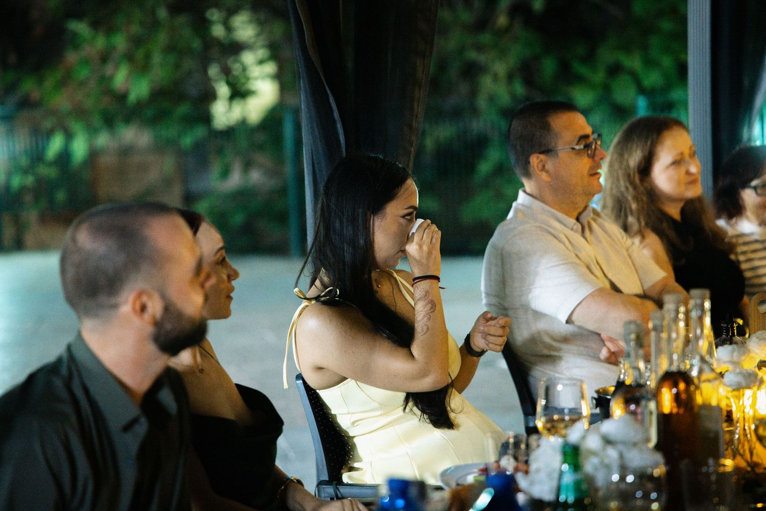 Wedding guests seated at outdoor evening reception table, woman wiping tears emotionally.