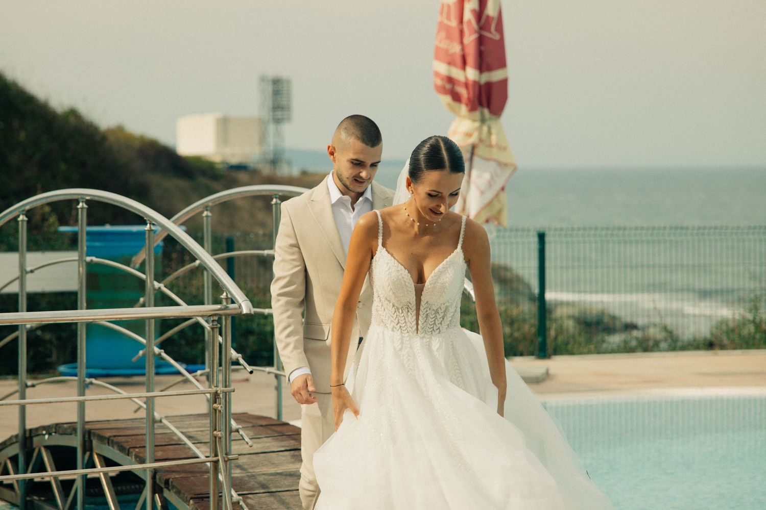 Bride and groom walk hand in hand by pool with ocean view.