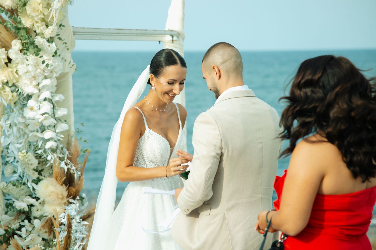 Smiling bride and groom hold hands during seaside wedding ceremony beside white flower arch with guest in red dress.