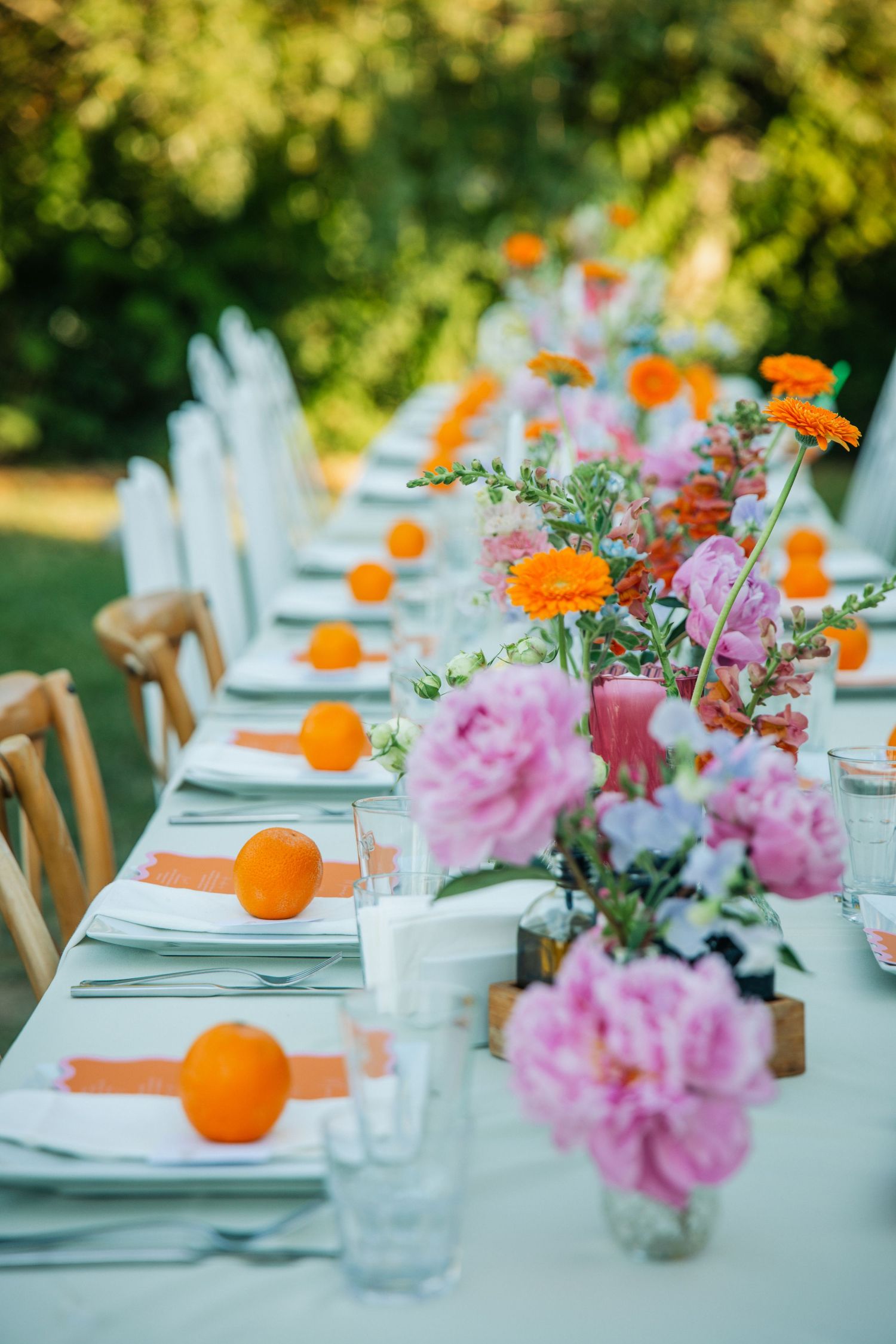 Elegant outdoor dining table with pink peonies, orange flowers, and fresh oranges as decor on a white tablecloth.