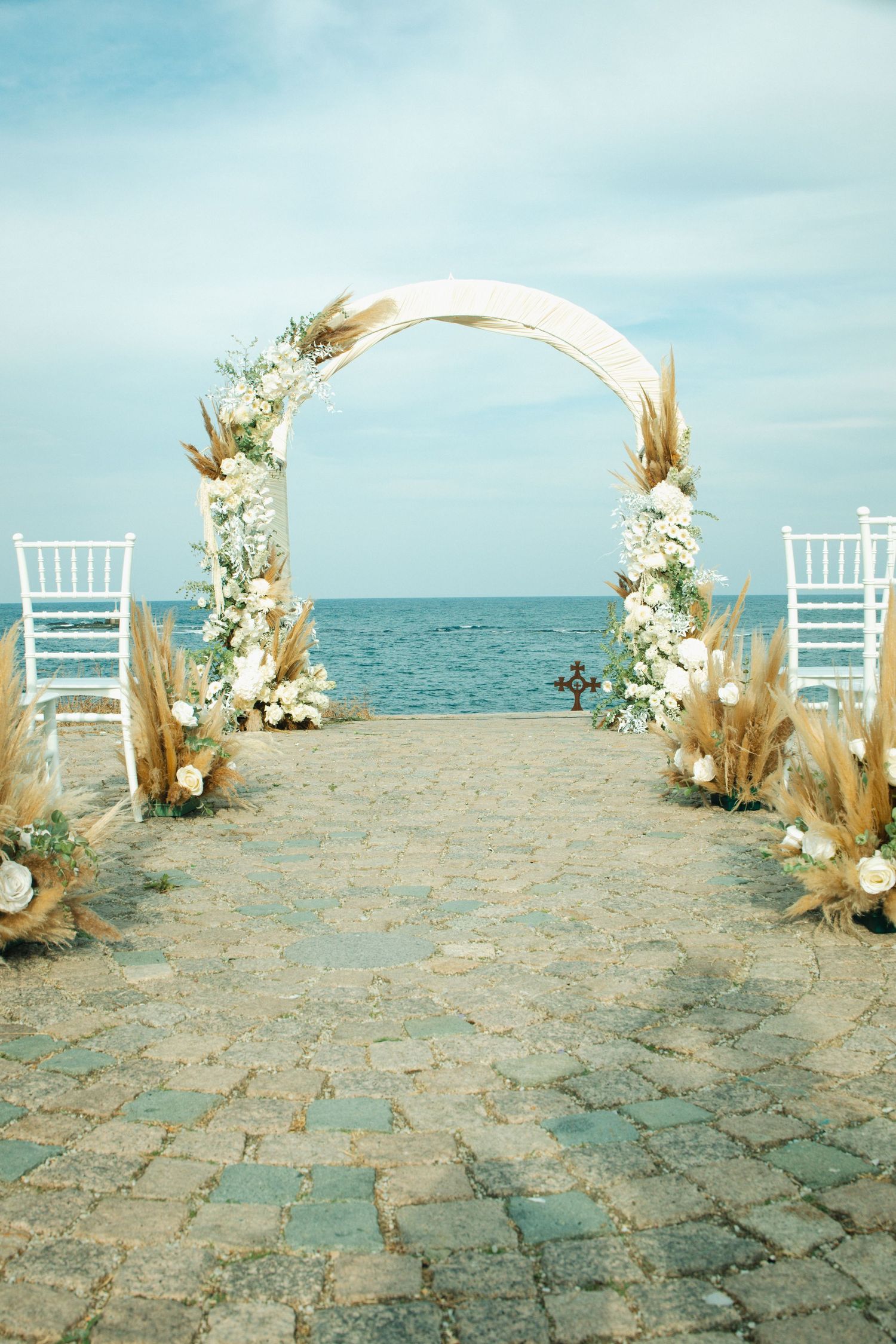 Elegant beach wedding arch adorned with white flowers and pampas grass, set on cobblestone with ocean views.