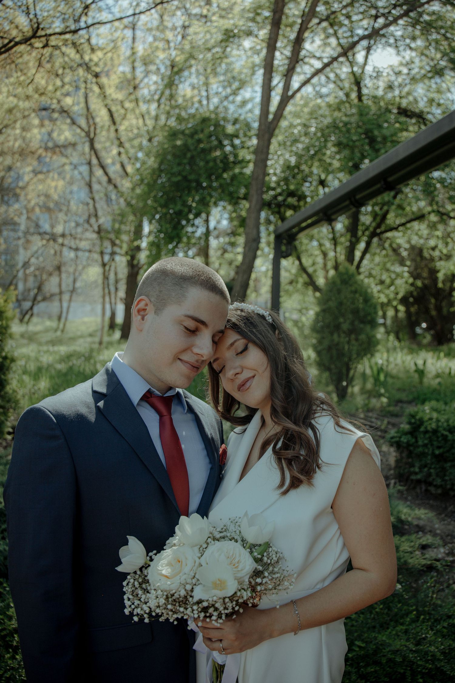 A newlywed couple embraces tenderly outdoors, bride holding white roses bouquet, groom in navy suit with red tie.