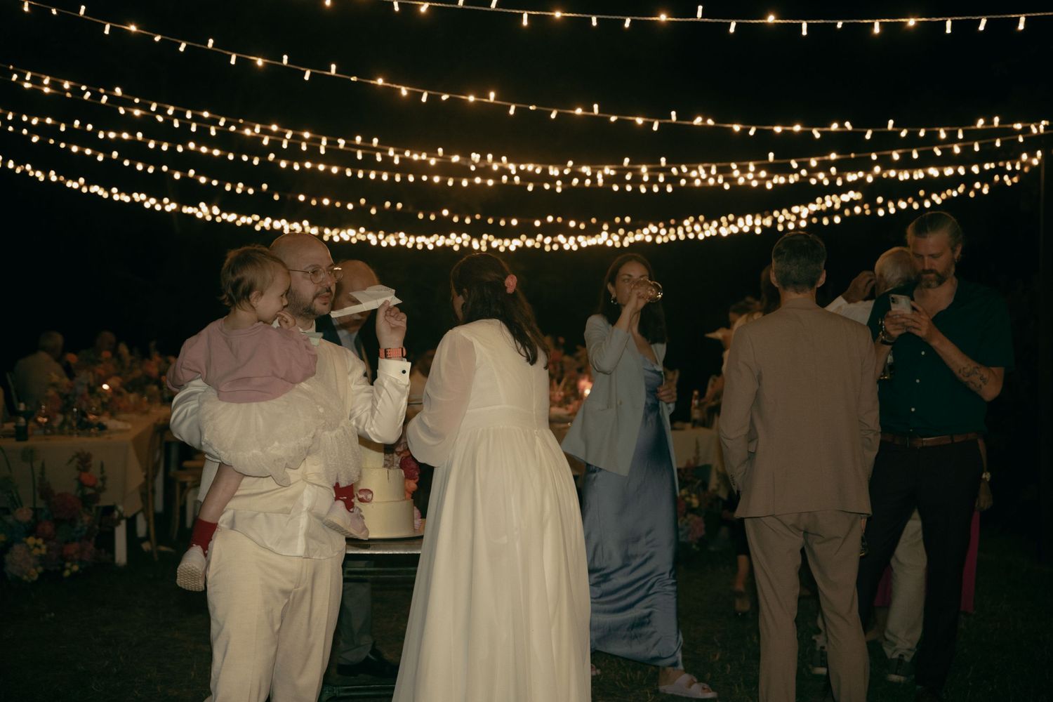 Bride and wedding guests dancing under canopy of glowing string lights at outdoor nighttime reception.