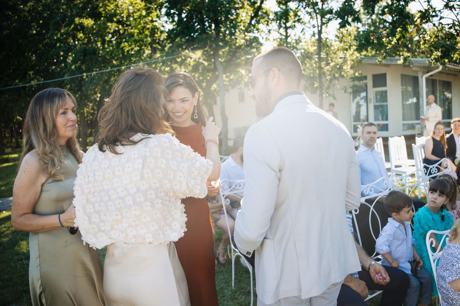 Wedding guests gather around groom in white suit during an intimate outdoor ceremony in golden sunlight.