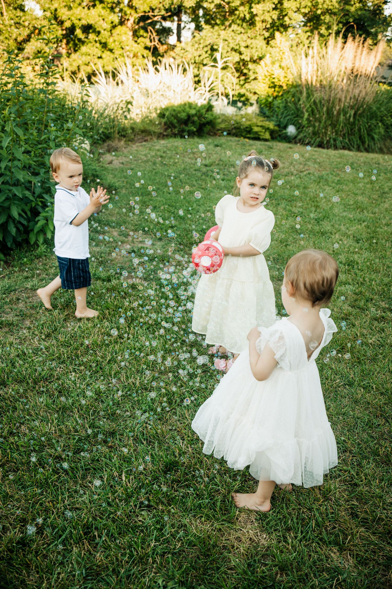 Three young children in white outfits play with soap bubbles on a green lawn at an outdoor celebration.