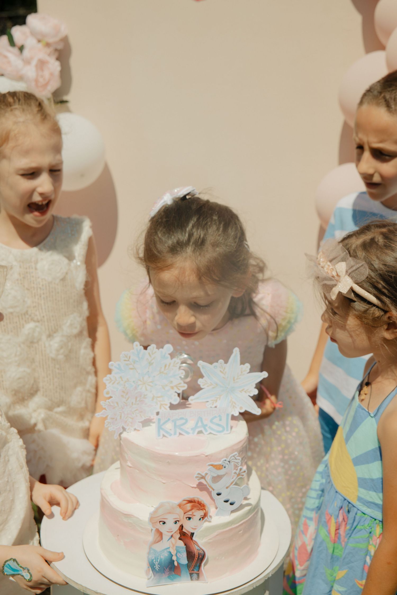 Girls gathered around a pink Frozen-themed birthday cake with Elsa decorations at a sunny outdoor party.
