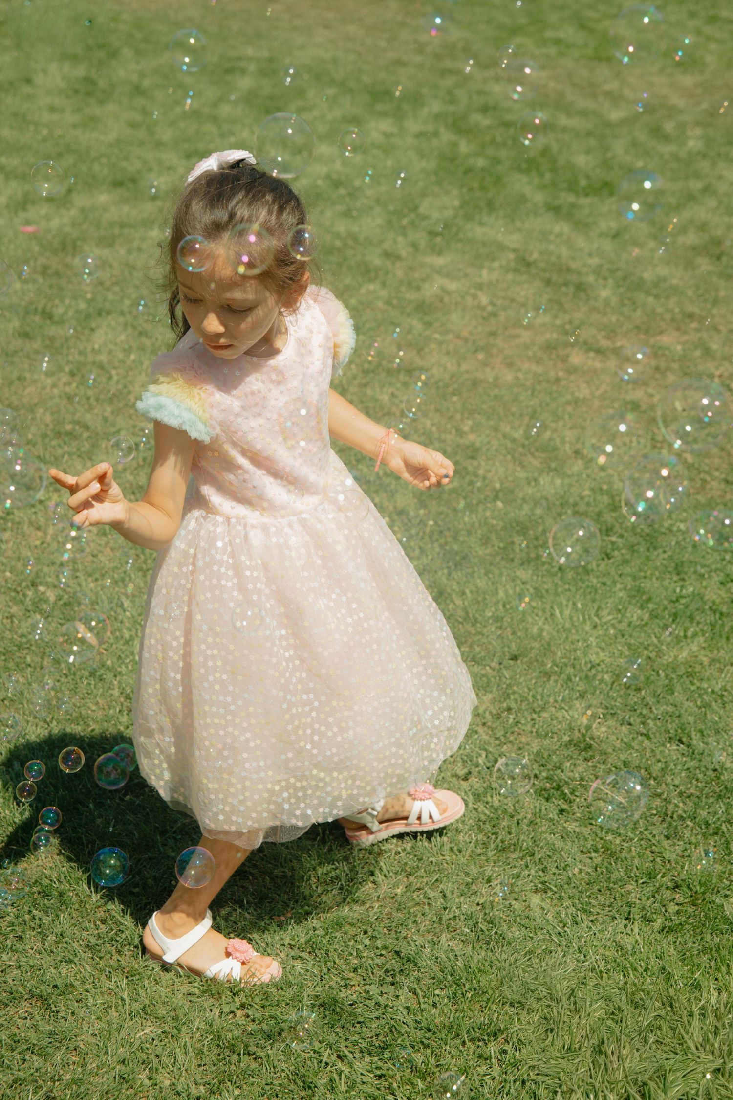 A young girl in a pink tulle dress and white sandals plays with bubbles on a sunny green lawn.