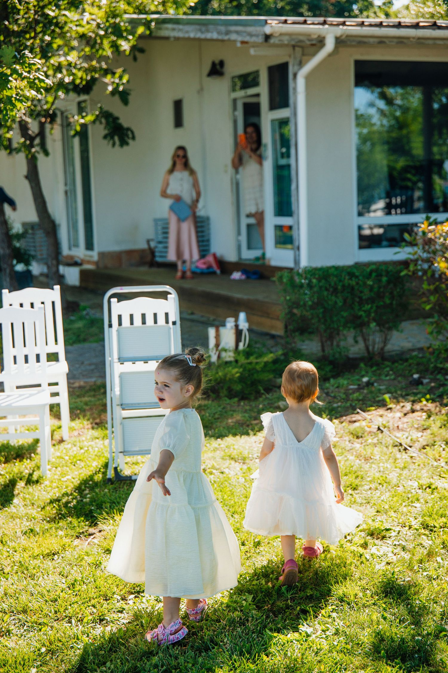 Two little girls in white dresses playing on a sunny garden lawn outside a white house.