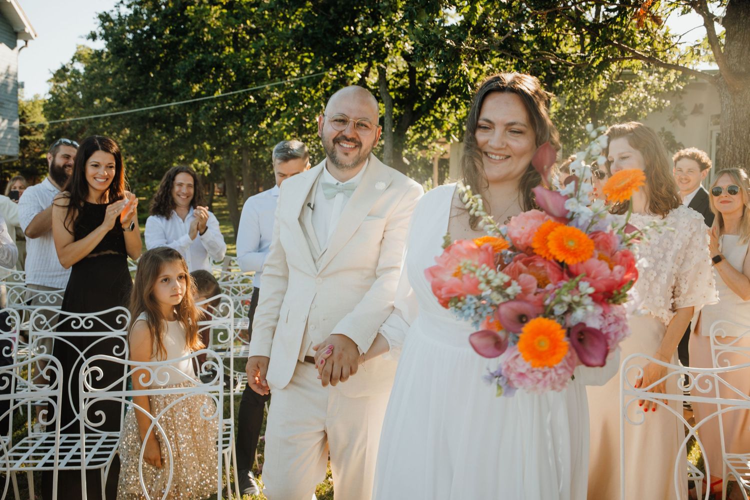 Newlywed couple walking down aisle holding colorful bouquet at outdoor garden wedding.
