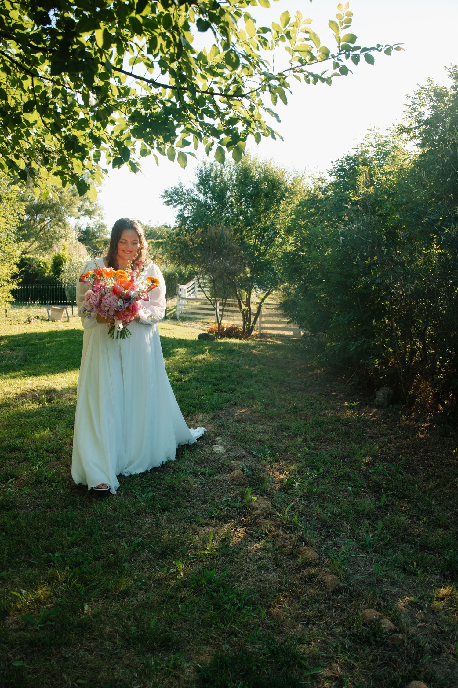 Bride in flowing white gown holds a vibrant bouquet of colorful flowers in a lush green garden.