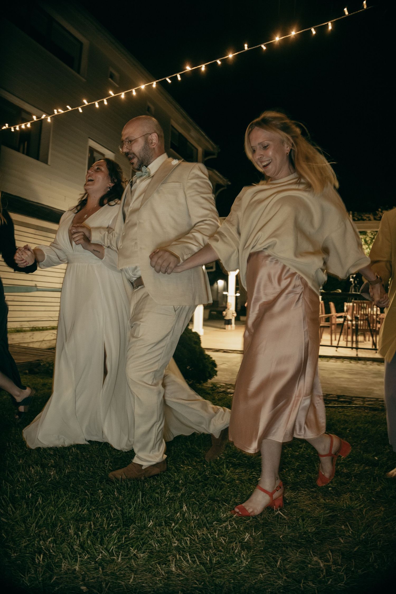 Bride, groom, and guest dancing outdoors at night under string lights.