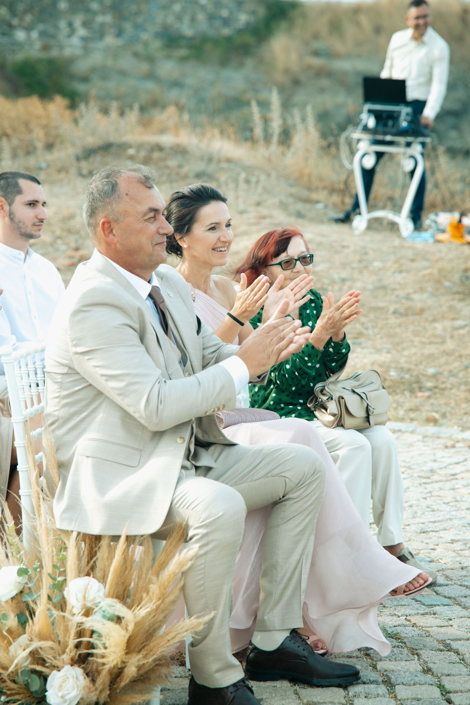 Wedding guests in formal attire applauding during outdoor ceremony with DJ equipment in background.