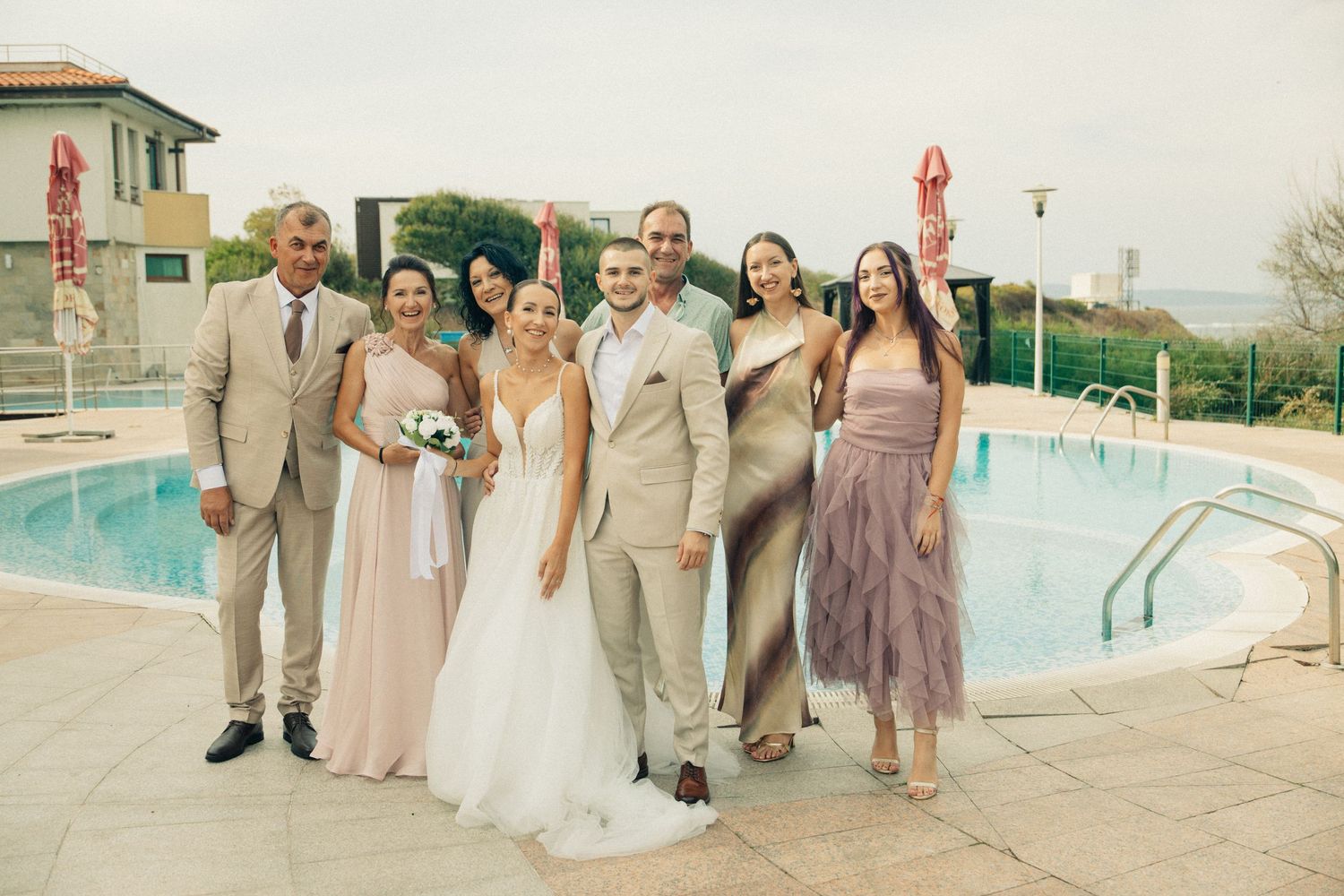 Wedding party group photo with bride and groom posing by outdoor pool at venue.
