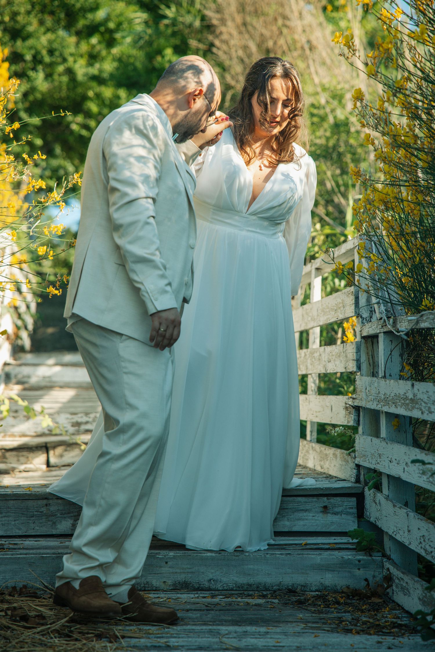 Bride and groom in white wedding attire share a tender moment on a rustic wooden bridge surrounded by yellow blooms.