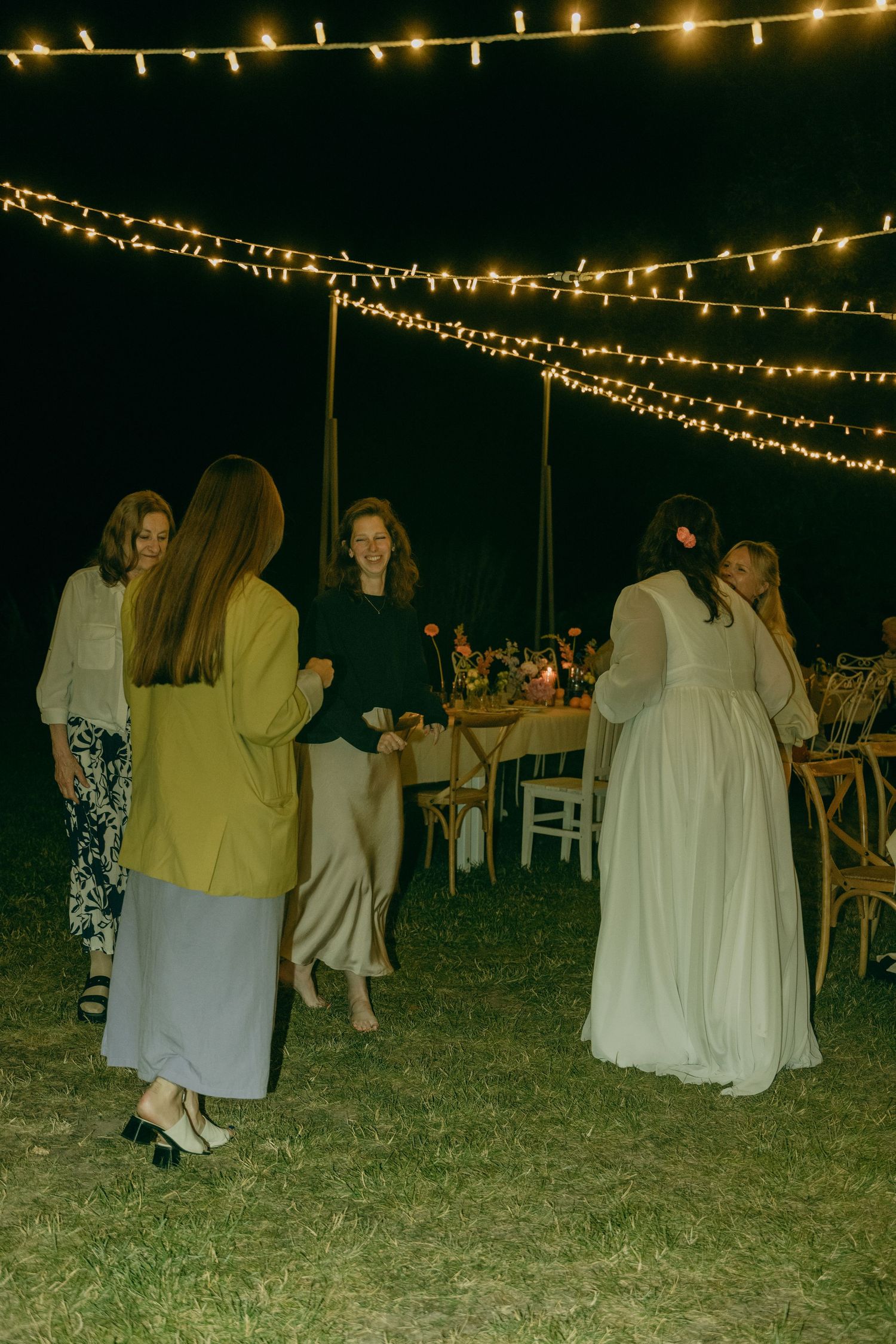 Bride in white gown mingles with guests under string lights at an outdoor evening wedding reception.