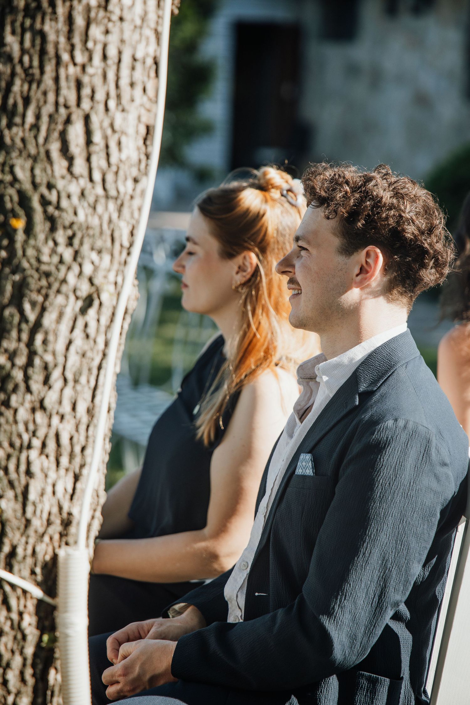 Smiling groom in gray suit sits beside bride during outdoor wedding ceremony in dappled sunlight.