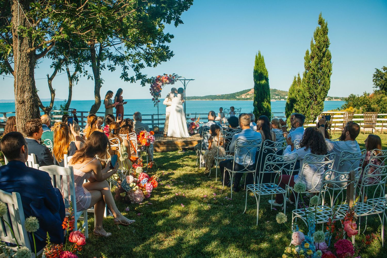 Outdoor waterfront wedding ceremony with guests seated on white chairs by the sea.