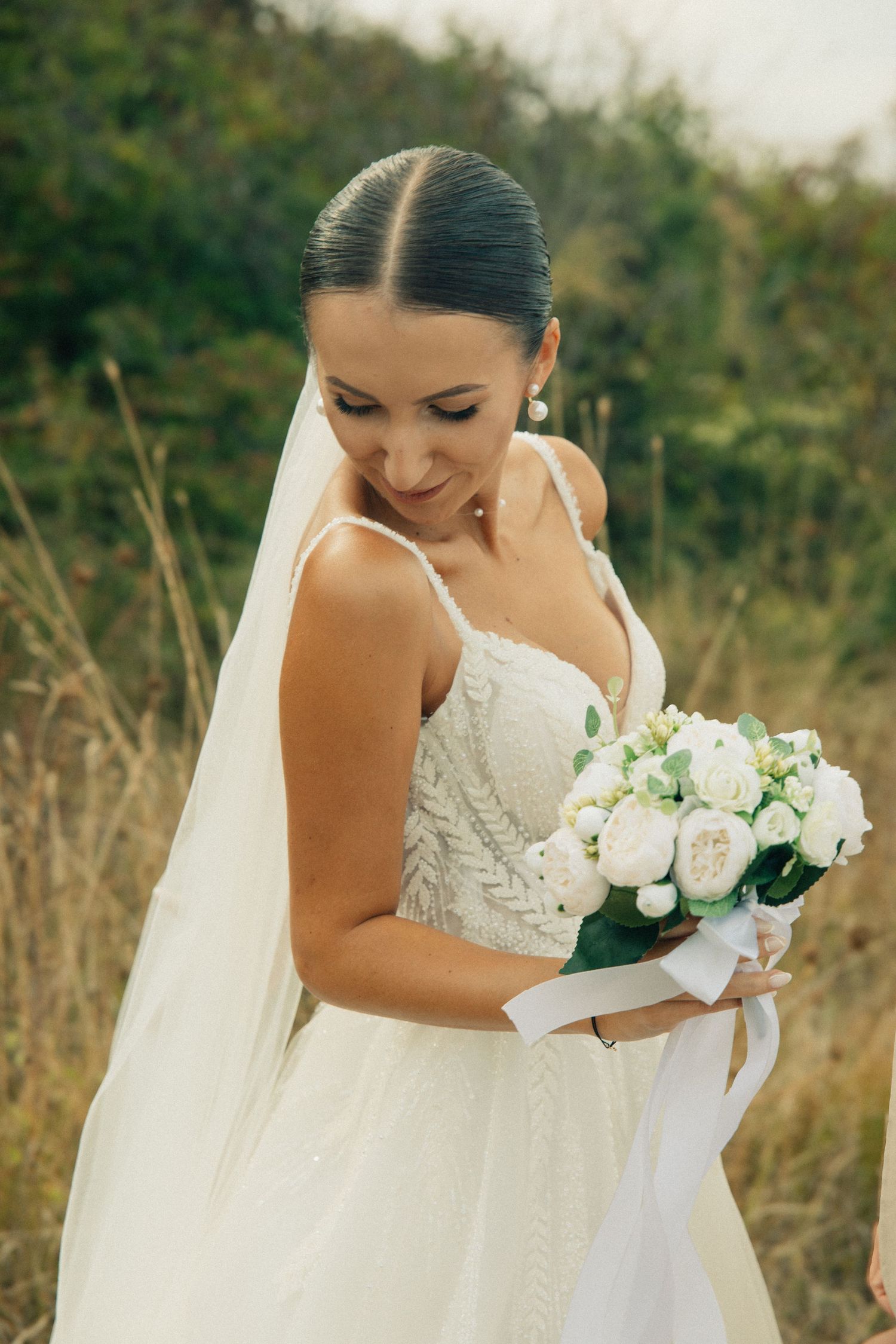 Smiling bride in white lace gown and veil holds bouquet of white peonies outdoors in natural setting.