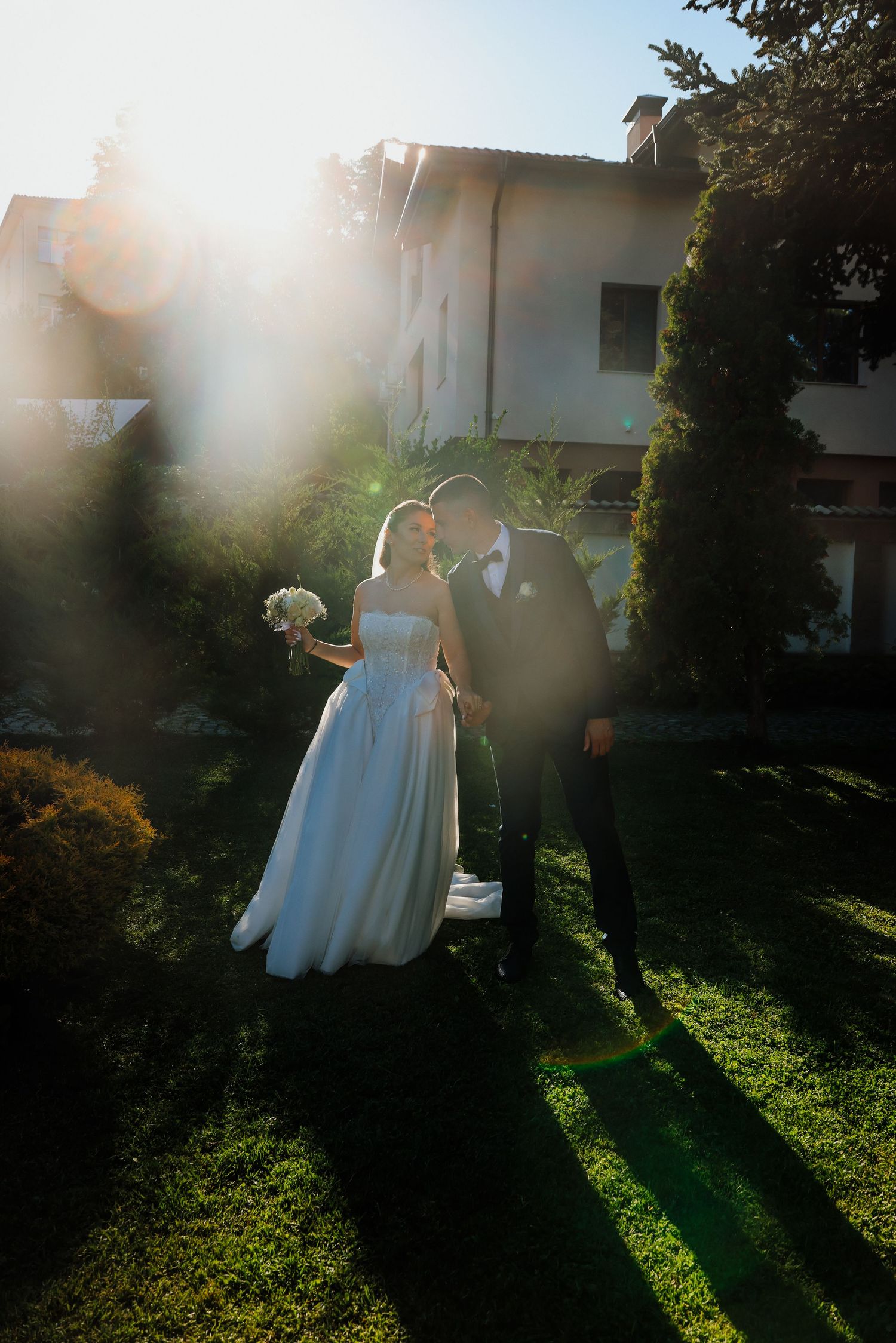 Bride and groom share a romantic kiss in a sunlit garden, bride holding white bouquet in elegant wedding dress.