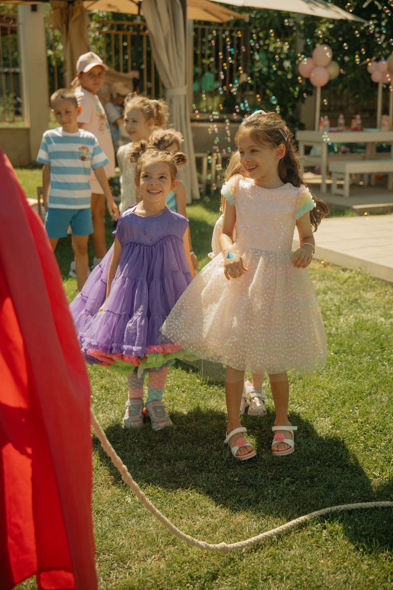 Two young girls in purple and white dresses smile at an outdoor birthday party on a sunny green lawn.