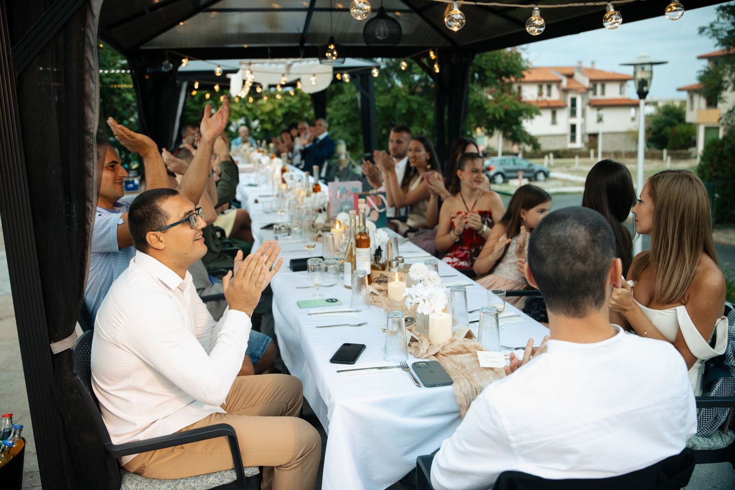 Large group of people enjoying an outdoor dinner party under a pergola with string lights at a long decorated table.