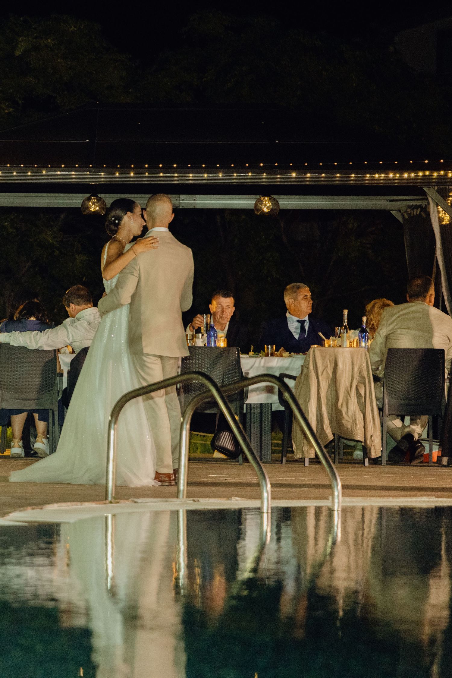 Bride and groom share first dance by illuminated poolside at night.