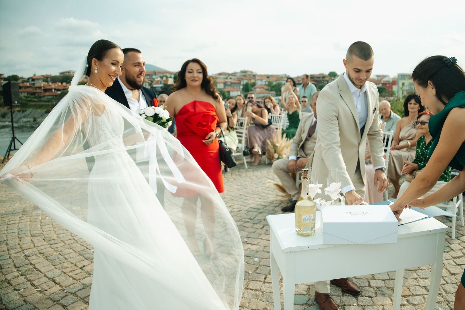 Bride and groom sign wedding documents outdoors with guests celebrating in background.