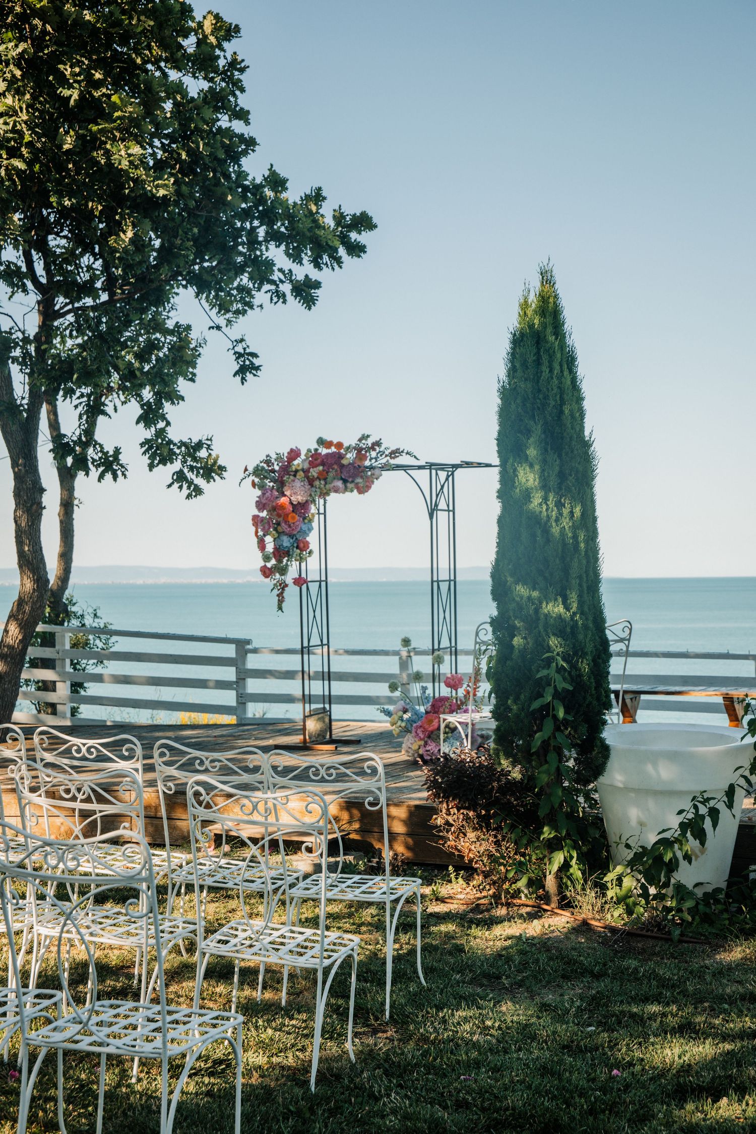 White wrought iron chairs arranged for an outdoor wedding ceremony with floral arch and ocean view.