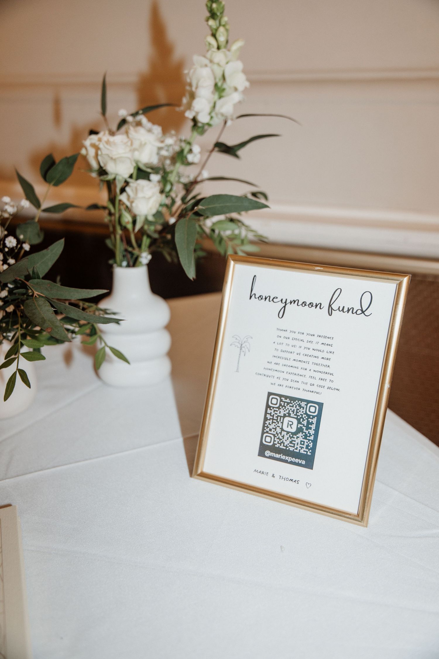 Gold-framed honeymoon fund sign with QR code beside white floral arrangement at wedding reception.