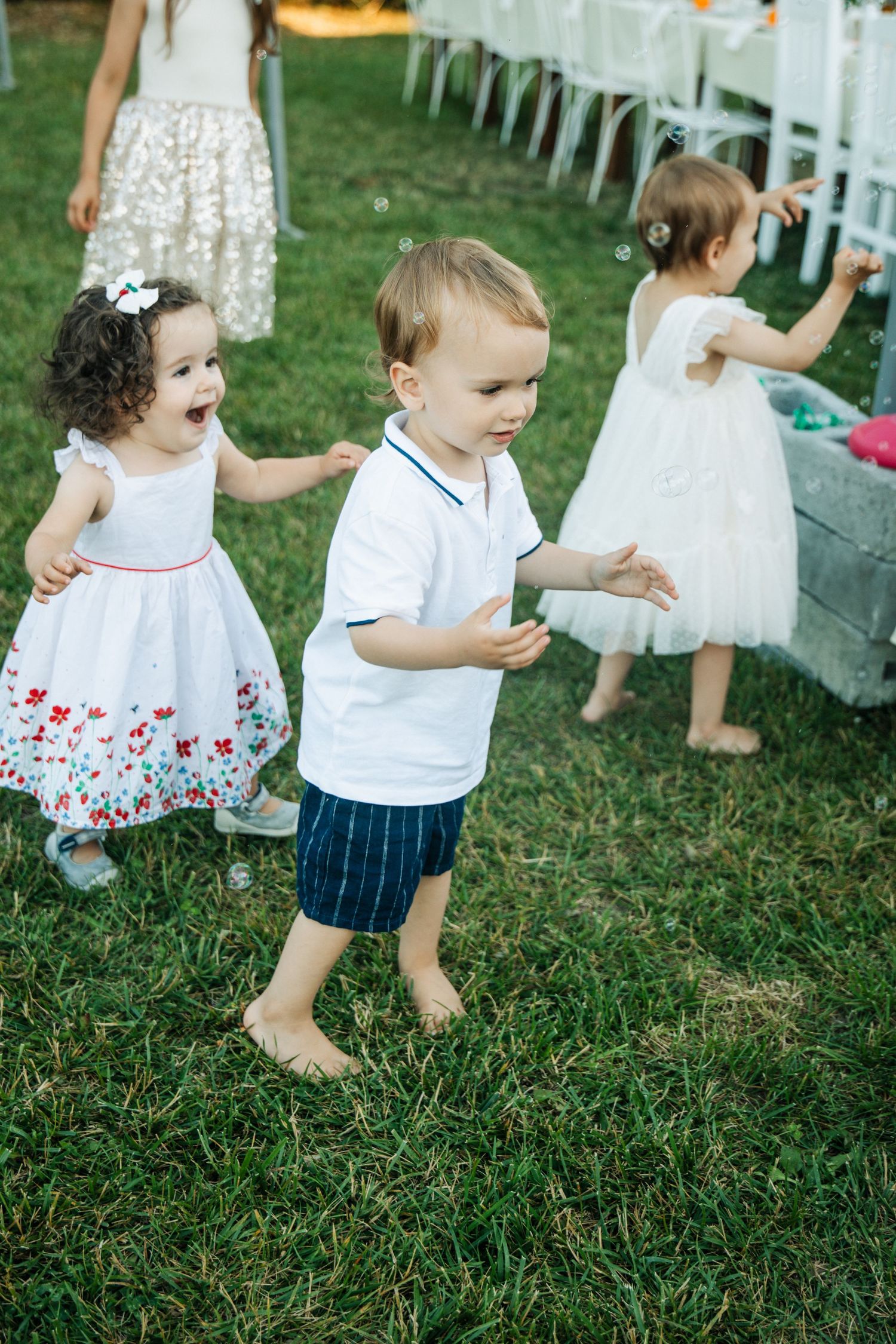 Three toddlers playing barefoot on green grass at outdoor wedding reception.