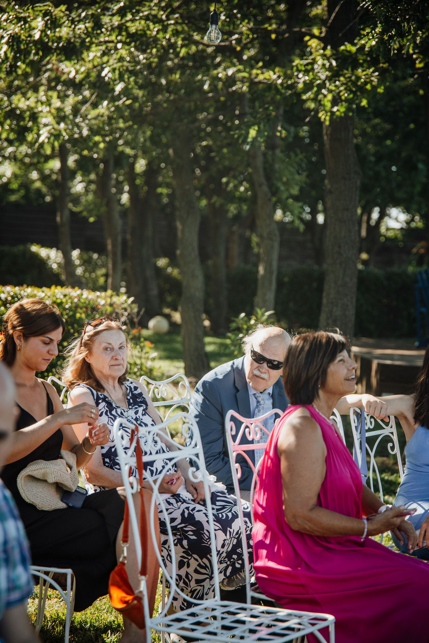 Guests seated on white ornate chairs at an outdoor wedding ceremony surrounded by lush green trees in sunlight.