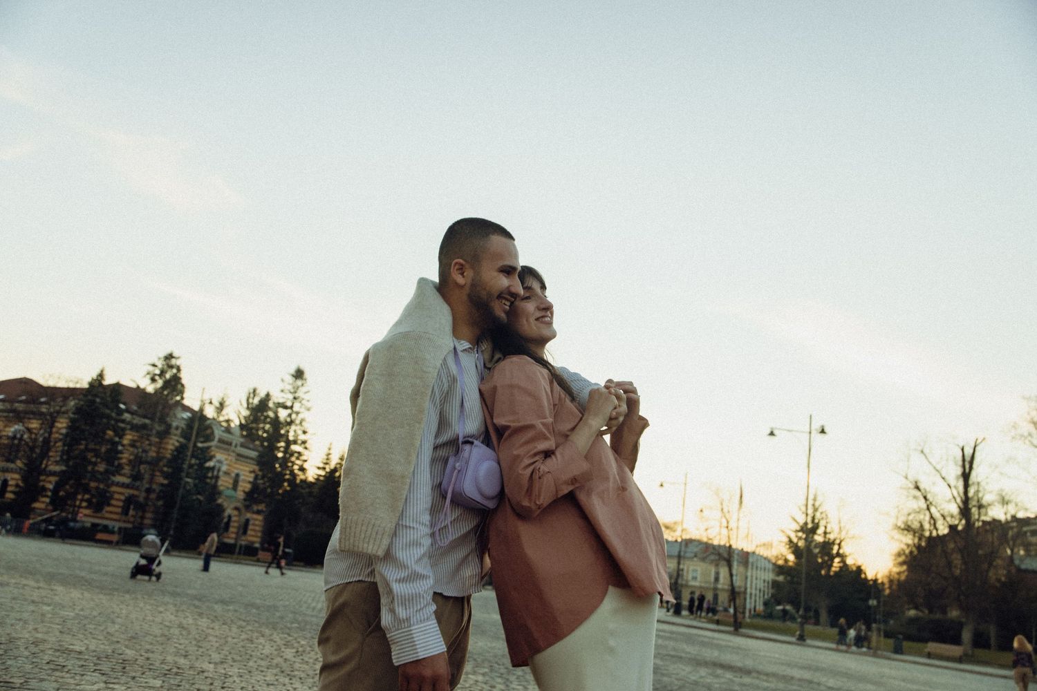 A romantic couple embraces during sunset in an outdoor setting with trees and buildings in the background.