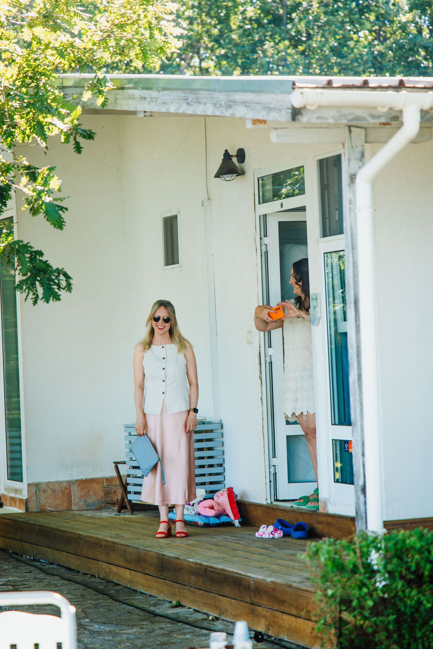 Woman in pink culottes and white top stands on porch of white house on sunny day.