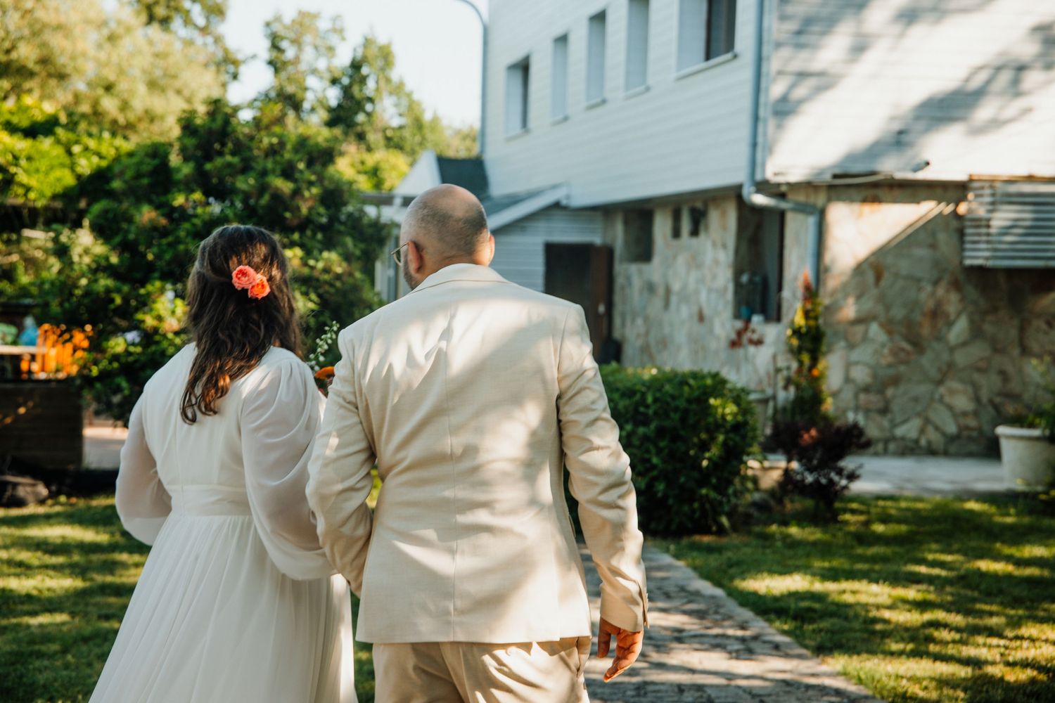 Bride and groom walking hand in hand through garden toward white stone building on their wedding day.