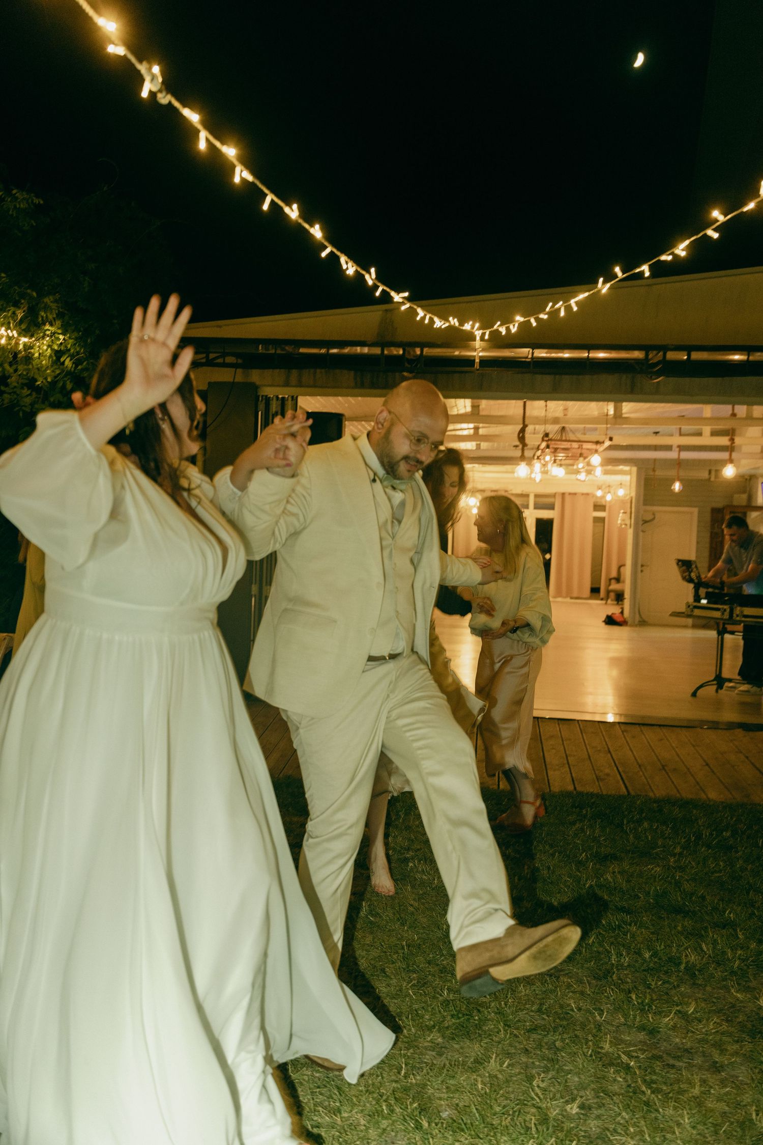 Joyful bride and bald groom dancing energetically outdoors under string lights at a nighttime wedding reception.