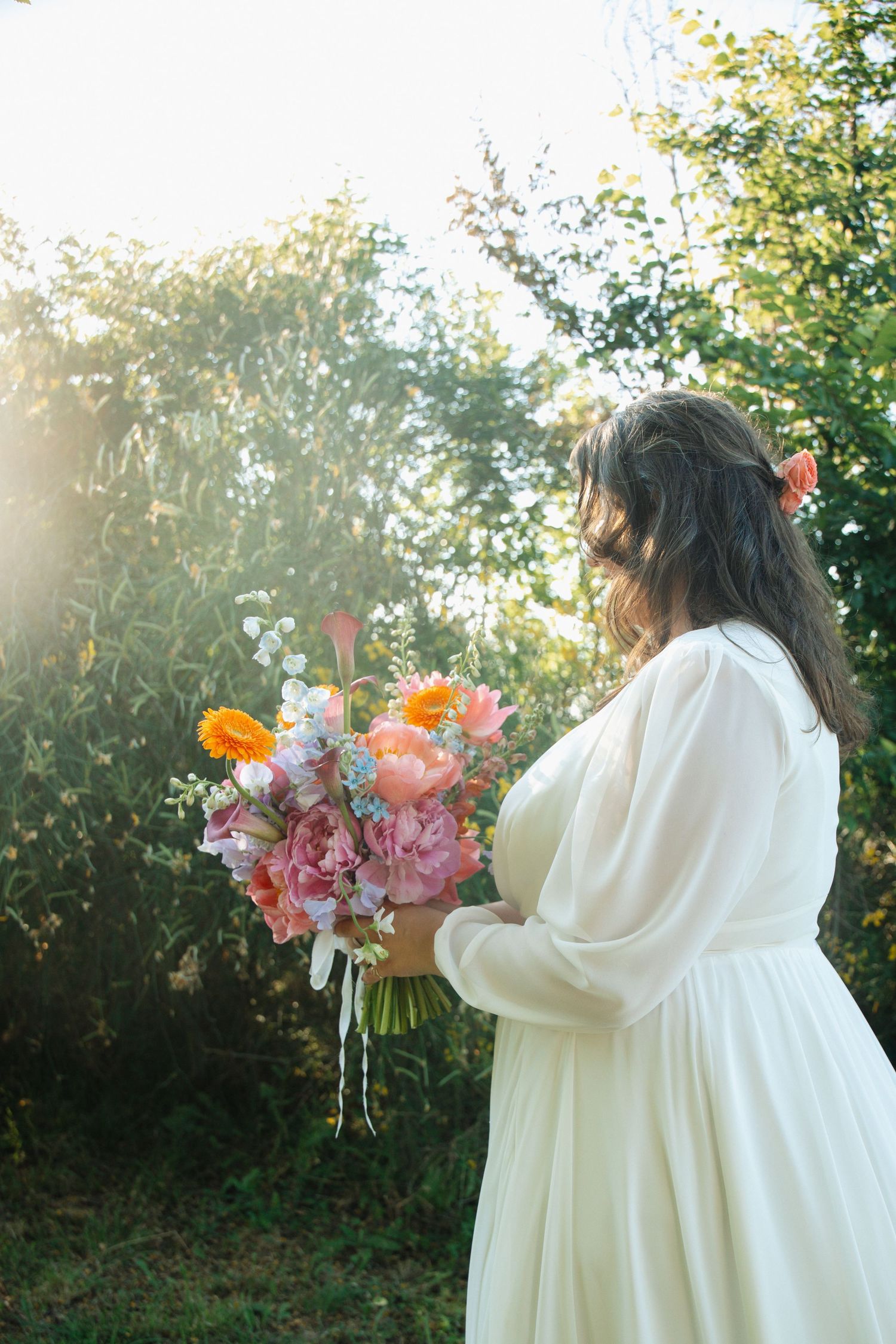 Bride in white gown holds a vibrant bouquet of pink, orange, and blue flowers in a sunlit garden setting.