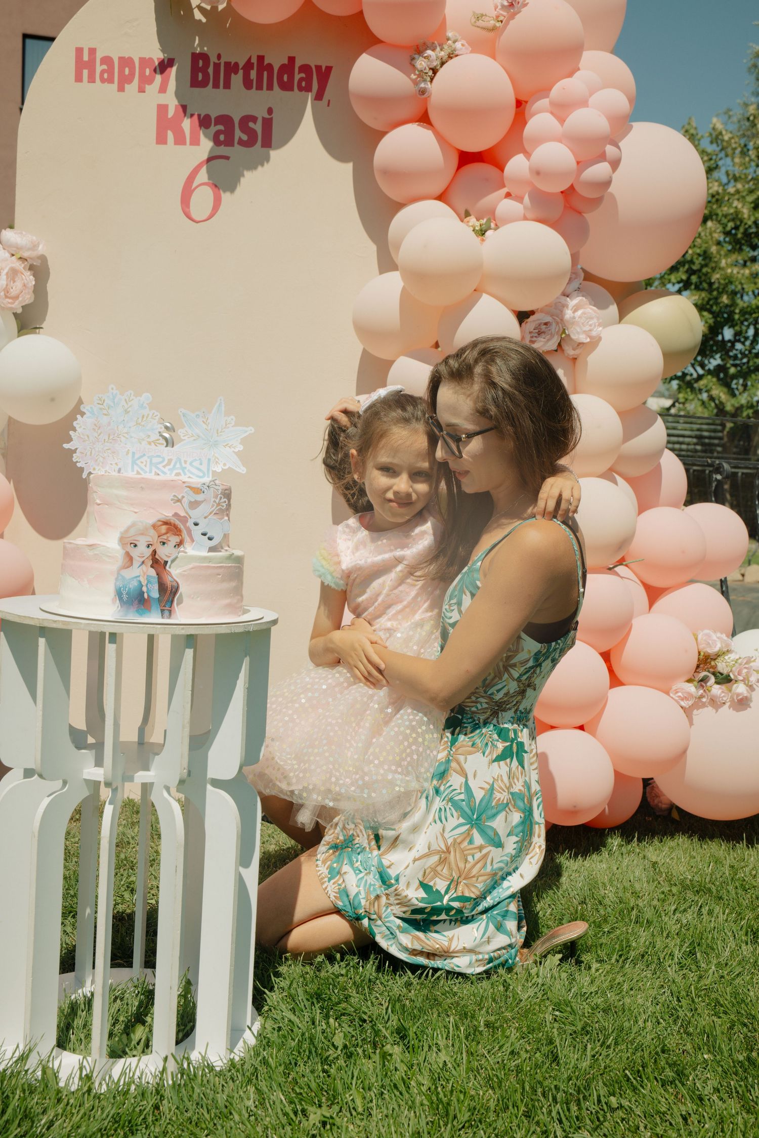 A mother hugs her daughter at a 6th birthday party with pink balloons and a Frozen-themed cake outdoors.