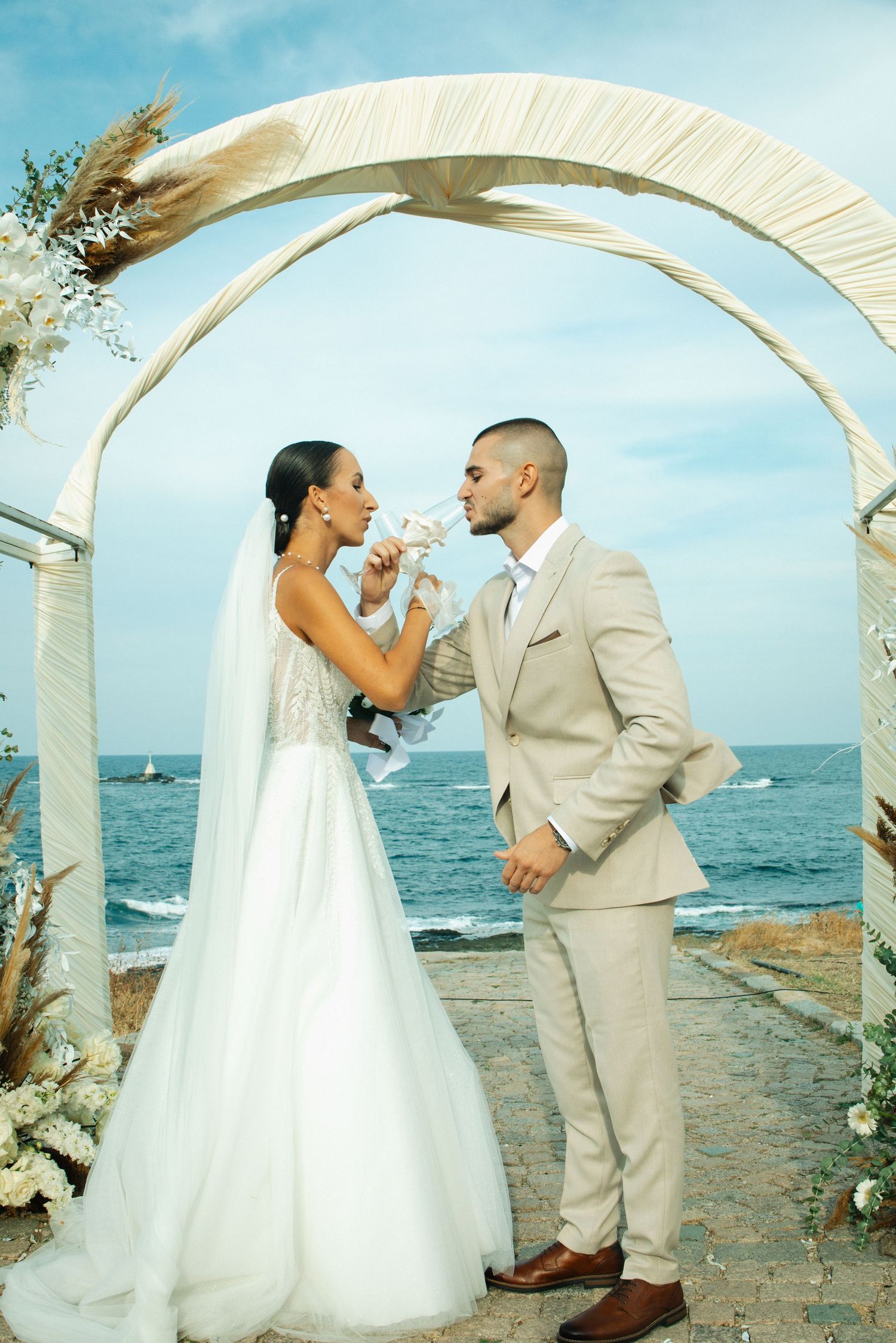 Bride and groom share a drink under floral arch by the sea.