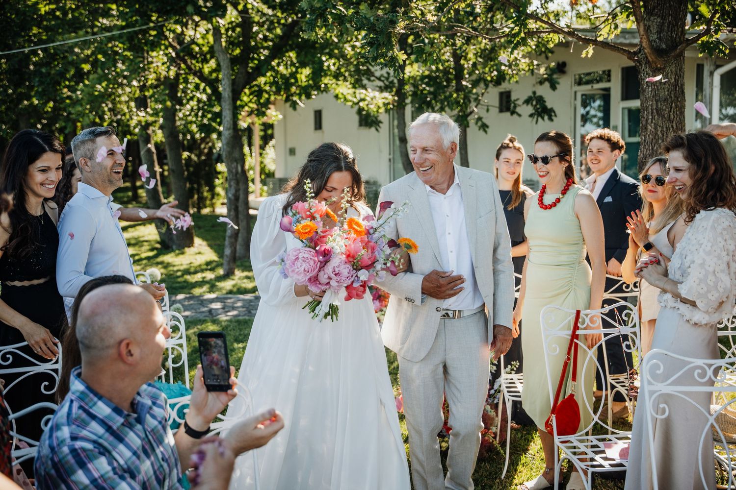 Bride in white gown holding colorful bouquet walks the aisle with older man at outdoor wedding.