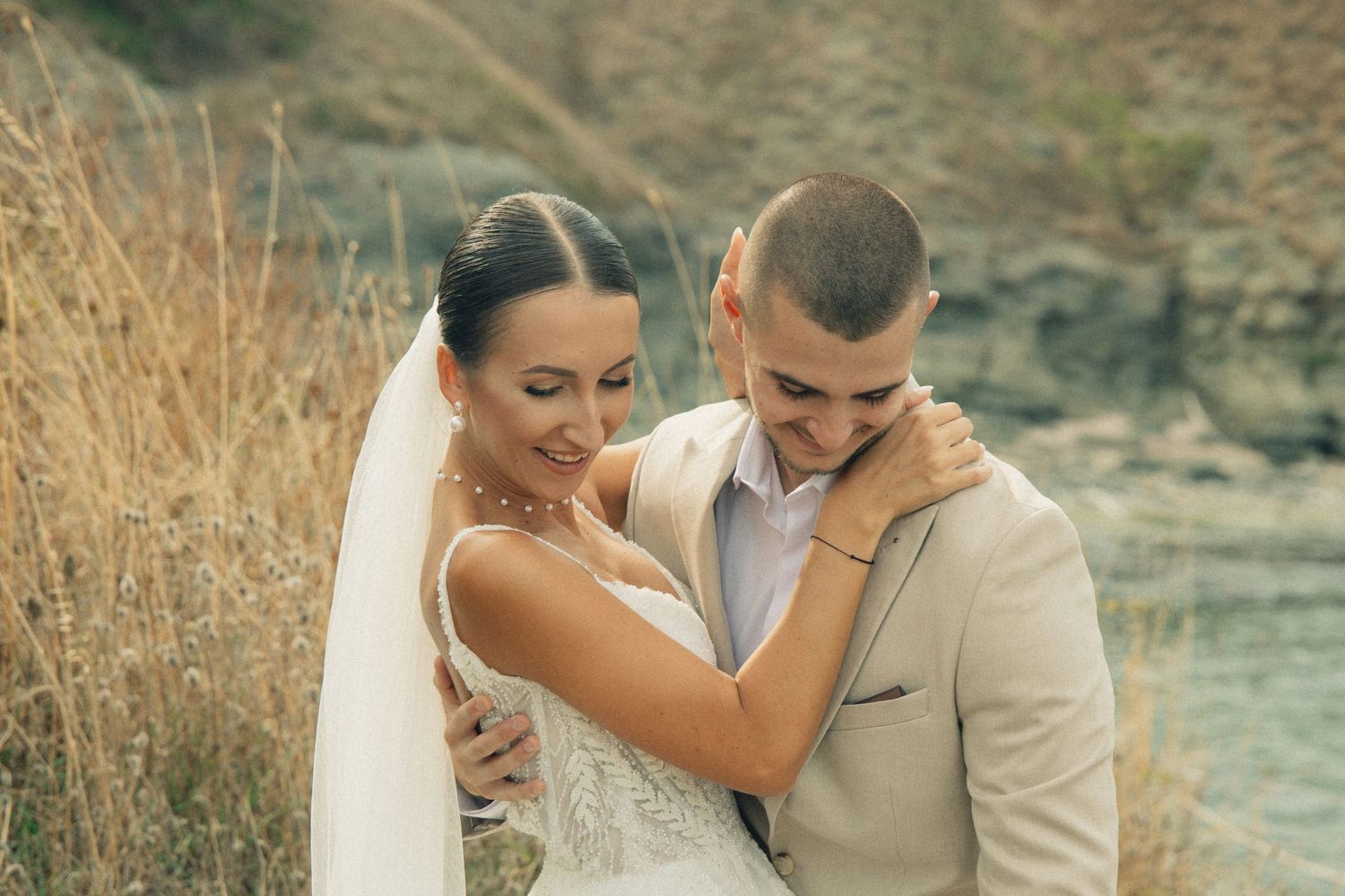 A bride and groom embrace outdoors near a river, wearing wedding attire with a veil and beige suit.