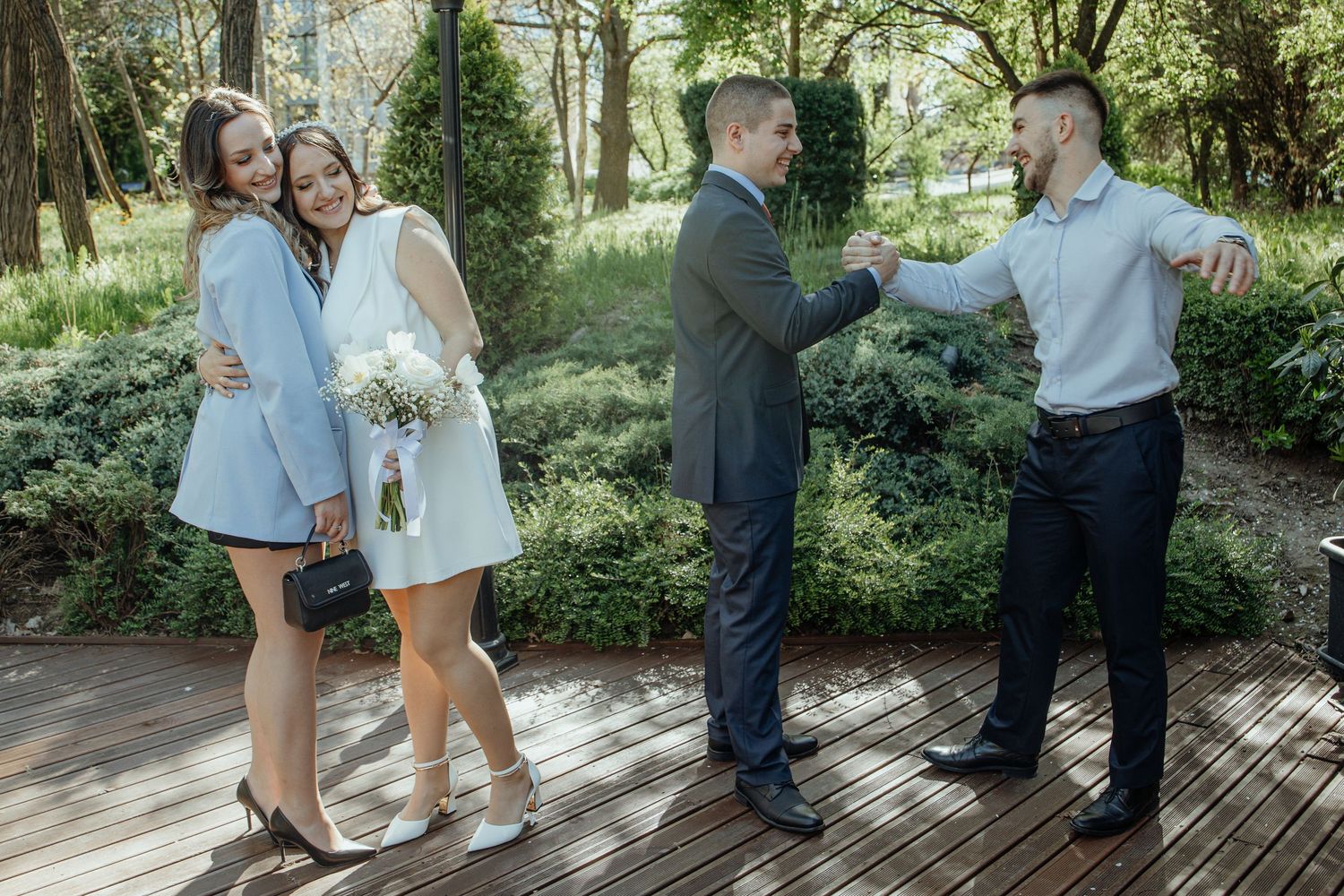 Couple in wedding attire poses in park while another man gestures nearby on wooden deck.