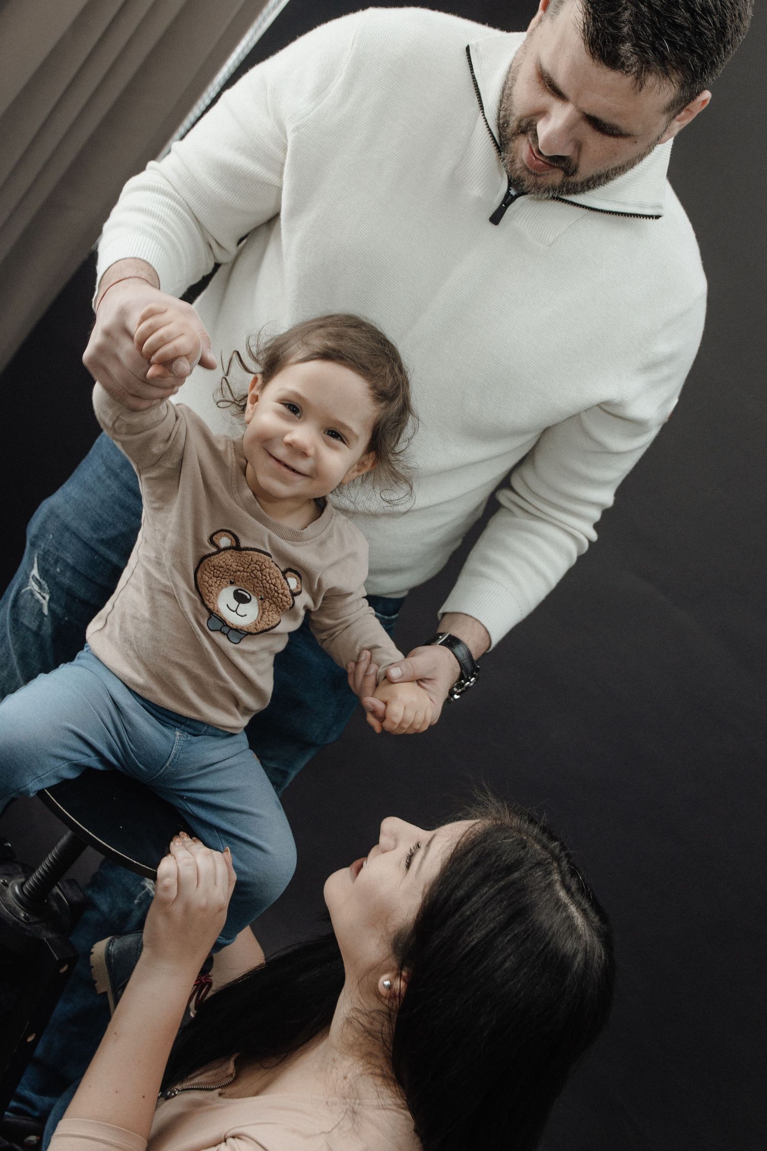 A happy family moment with a man in white sweater lifting a smiling toddler in bear shirt while woman looks up lovingly.