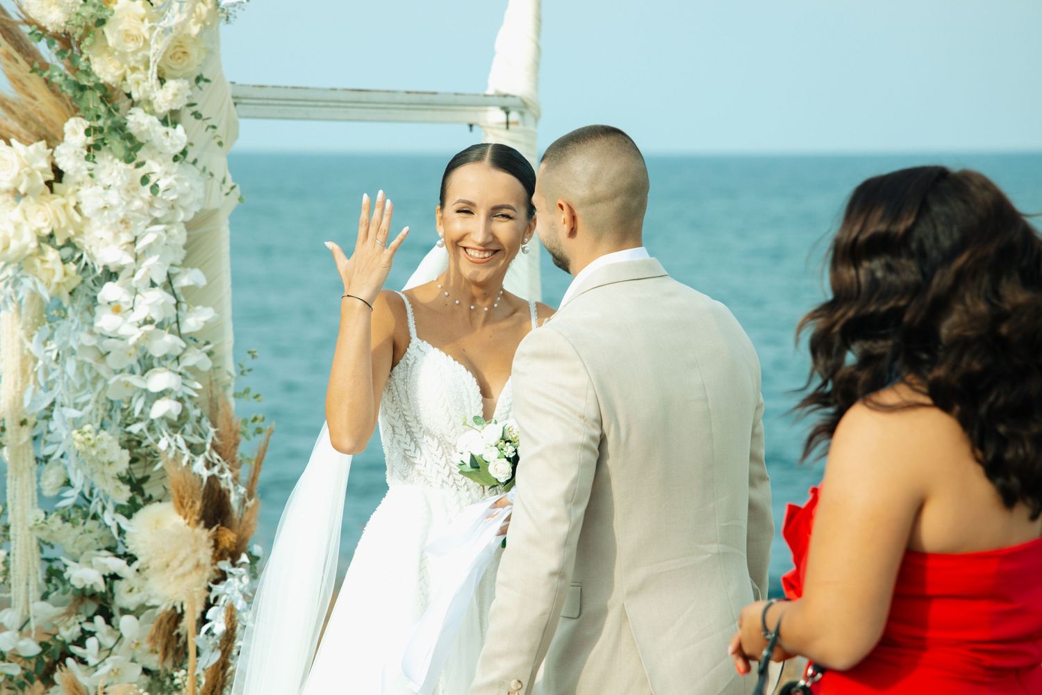 Bride in white gown waves during outdoor beach wedding ceremony with groom and bridesmaid in red dress.