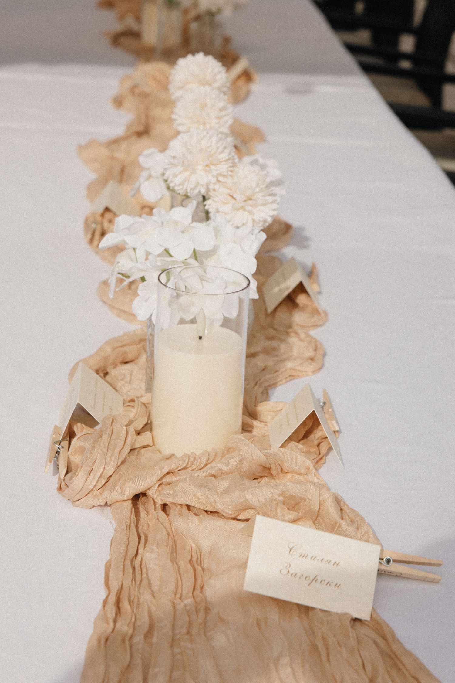 Elegant wedding table runner with white flowers, candles and place cards on draped beige fabric.