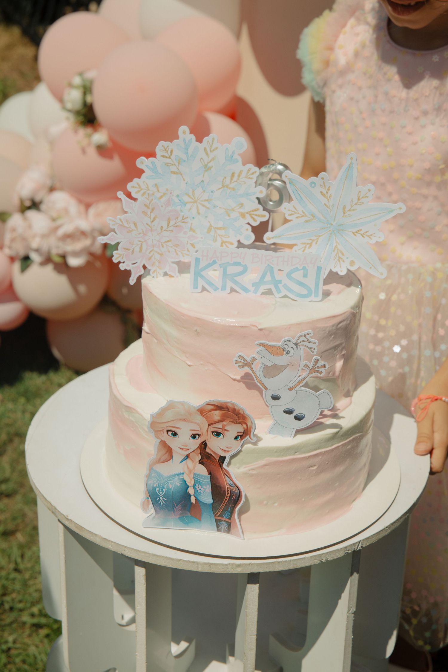 Two-tier Frozen themed birthday cake with Elsa, Anna, Olaf, and snowflake topper reading KRASI on white stand.