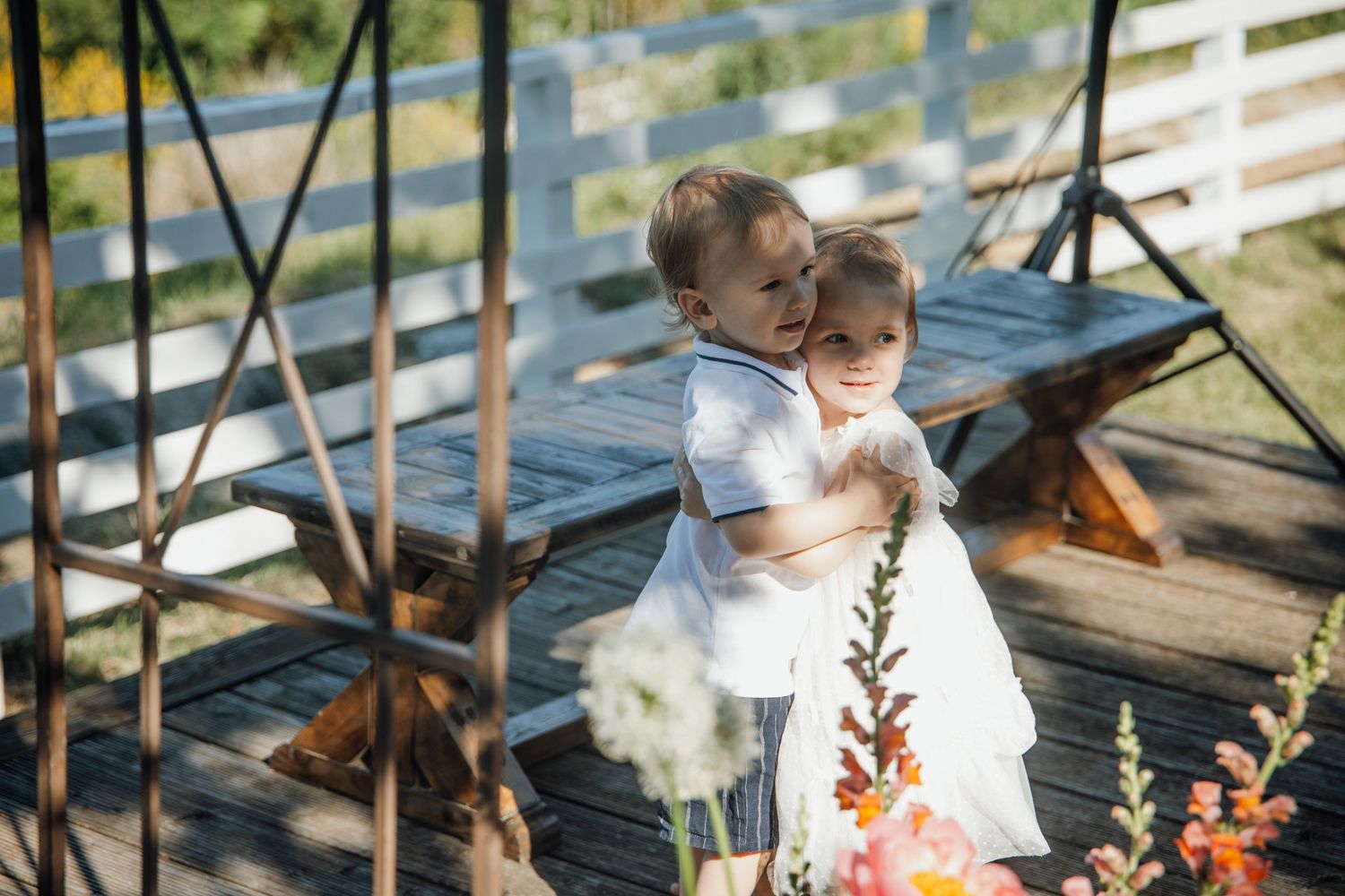 Two young children in white outfits share a sweet hug on a wooden deck at an outdoor wedding celebration.