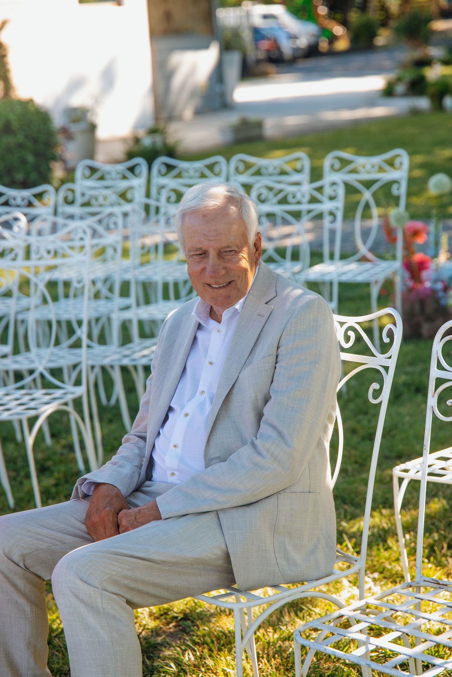 Elderly man in beige suit sitting on white ornate chair at outdoor wedding ceremony.