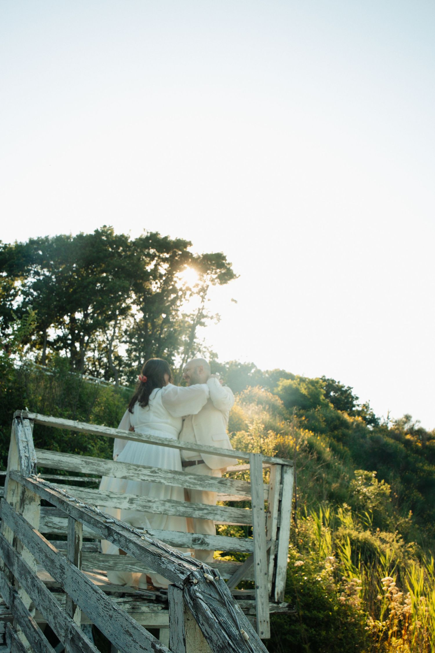 Two women in white dresses stand on rustic wooden stairs surrounded by lush greenery during golden hour sunset.
