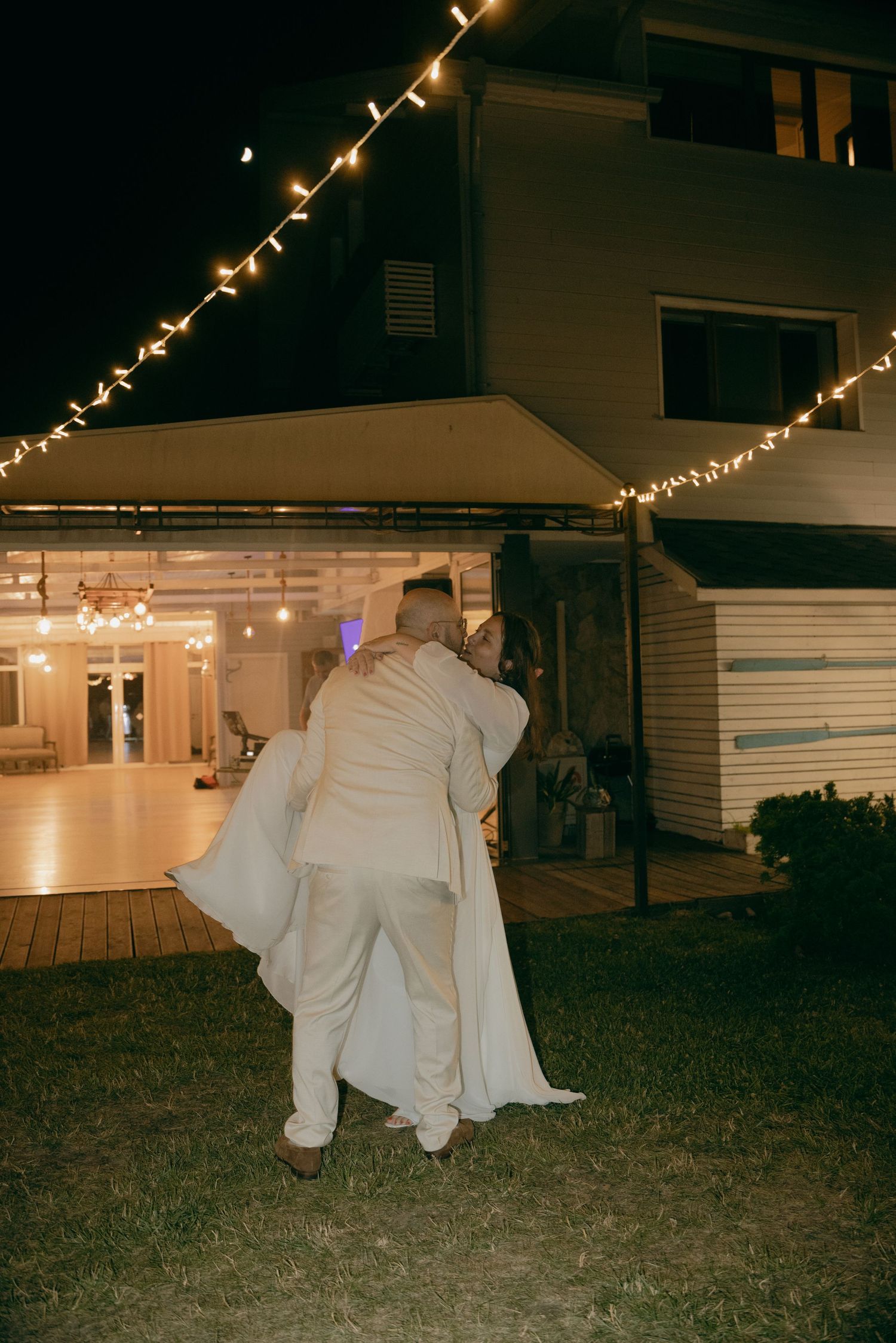 Groom carrying bride in white dress outside illuminated venue at night with string lights.